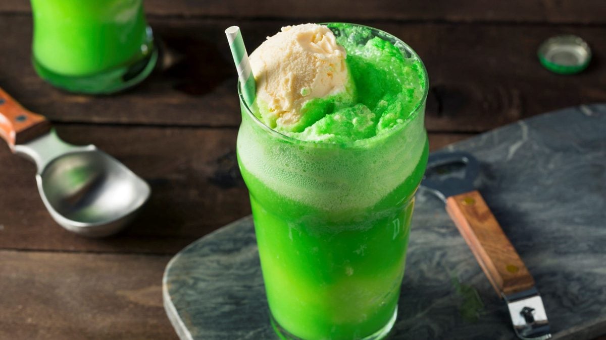 St. Patrick's Day Green Soda Float im Glas mit Strohhalm auf grauer Platte. Daneben Löffel mit Holzgriff. Links Eislöffel und ein weiteres Glas angedeutet. Frontalaufnahme.