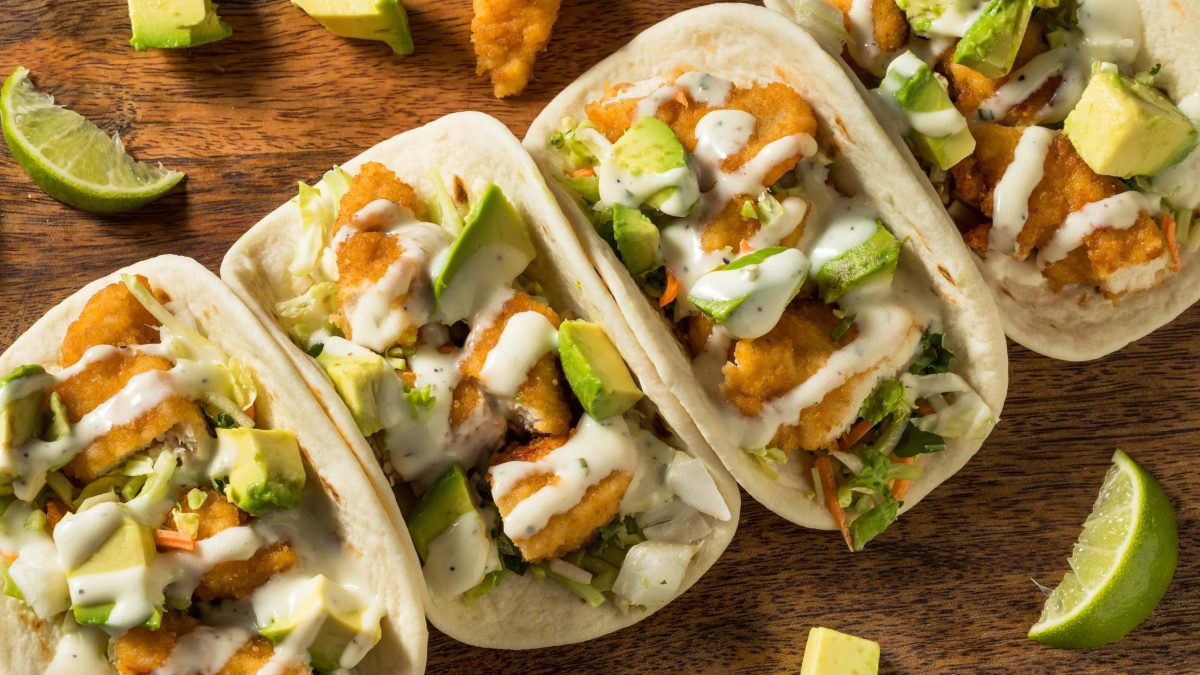 Fried Chicken Tacos von oben mit paniertem Hähnchen und Avocado Stücken auf Holztisch.