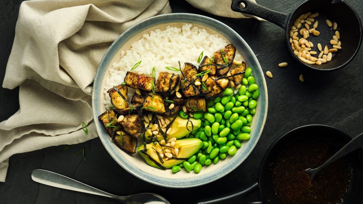 Teriyaki-Bowl mit Aubergine, Edamame, Avocado, Reis und Pinienkernen auf dunklem Untergrund in der Draufsicht, daneben ein Geschirrtuch und eine Schale Pinienkerne.