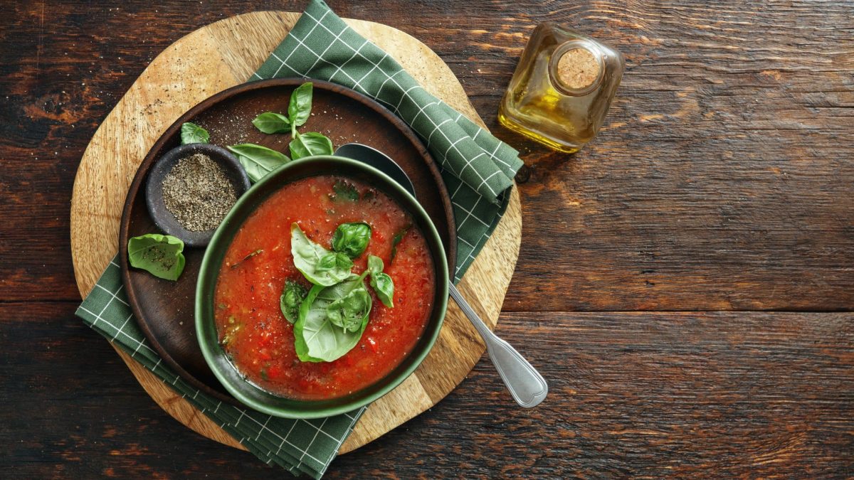 Eine Schüssel Tomaten-Sellerie-Suppe mit Basilikum auf Holz in der Draufsicht, daneben steht eine Flasche Öl.