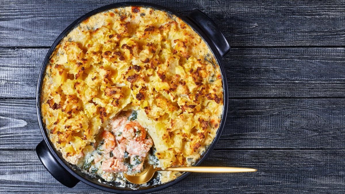 Mit Kartoffelbrei überbackene Lachspfanne mit Spinat und Sahne in einer großen Form auf einem Holztisch.