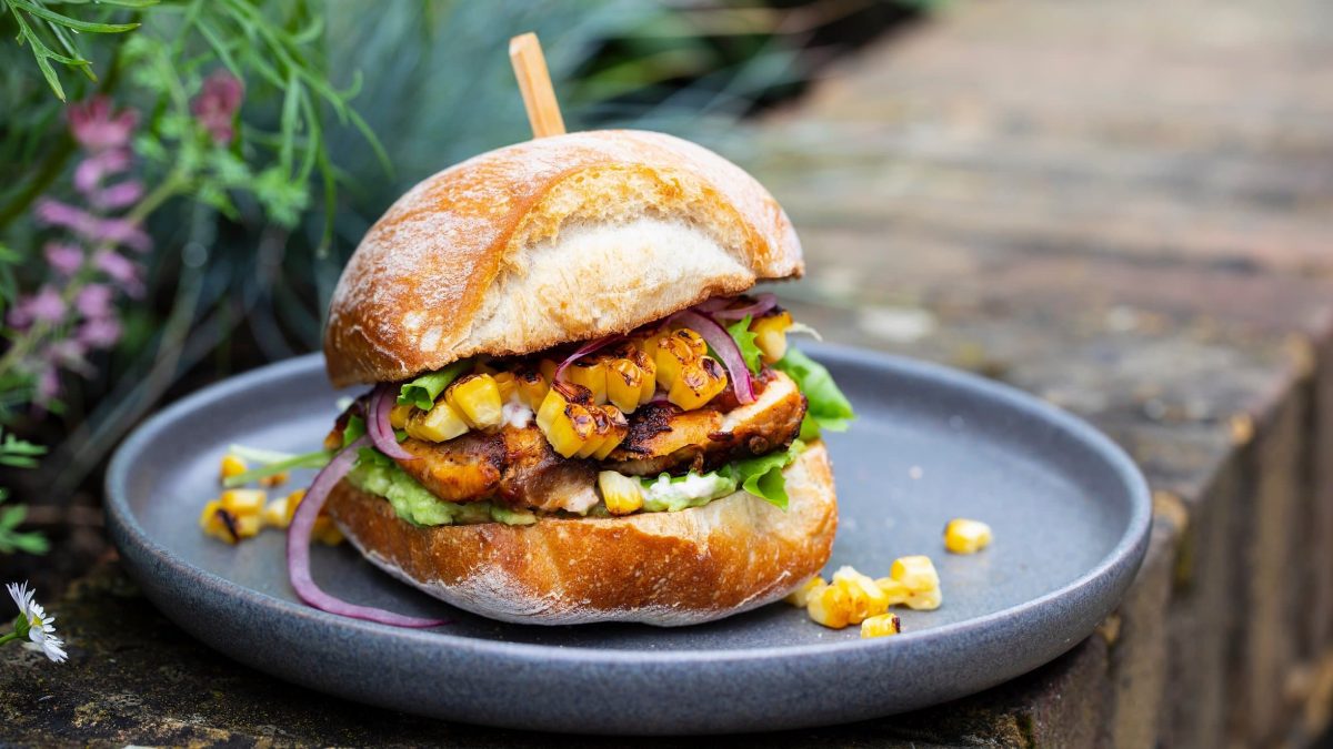Ein veganer Elote-Burger mit Mais, Salat und selbst gemachtem Bratling auf einem grauen Teller.