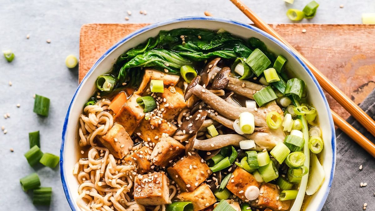 Eine Schale vegane Miso-Ramen mit Räuchertofu, Frühlingszwiebeln, Pilzen, Nudeln, Pak Choi und Sesam in der Draufsicht, daneben liegen Essstäbchen.