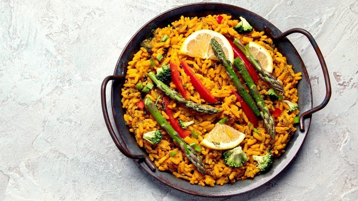 Eine gusseiserne Pfanne mit veganer Paella mit grünem Spargel, Paprika, Brokkoli und Zitronenscheiben.