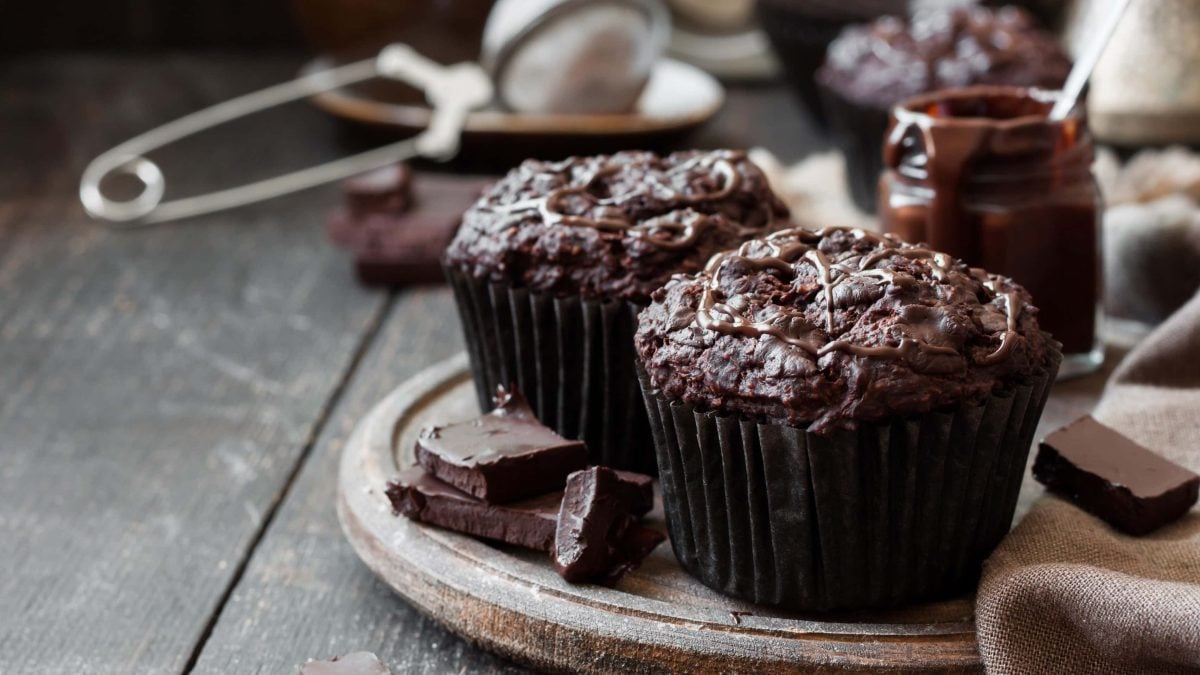 Zwei vegane Schoko-Himbeer-Muffins auf einem runden Holzbrett, daneben liegen Kuvertüre-Stücke, im Hintergrund sind unscharf Küchenutensilien zu sehen.