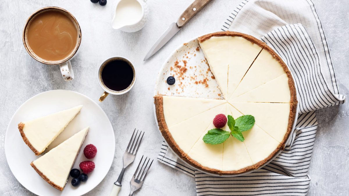 Ein angeschnittener veganer New York Cheesecake in der Draufsicht, daneben steht ein Teller mit zwei Stück Kuchen sowie zwei Kaffeetassen und ein Milchkännchen.