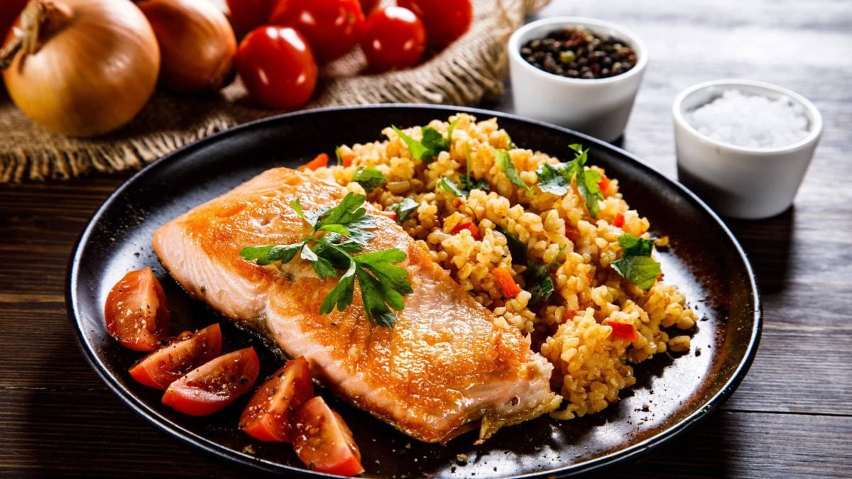 Gedämpfter Wildlachs mit Safran-Couscous und geschmolzenen Tomaten auf schwarzem Teller und grauem Tisch. Im Hintergrund Tomaten und Zwiebel auf Holztablett. Zwei Schalen mit Pfeffer und Salz rechts im Bild. Frontalansicht.