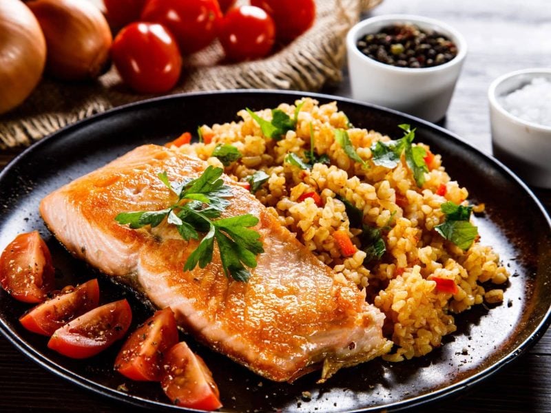 Gedämpfter Wildlachs mit Safran-Couscous und geschmolzenen Tomaten auf schwarzem Teller und grauem Tisch. Im Hintergrund Tomaten und Zwiebel auf Holztablett. Zwei Schalen mit Pfeffer und Salz rechts im Bild. Frontalansicht.