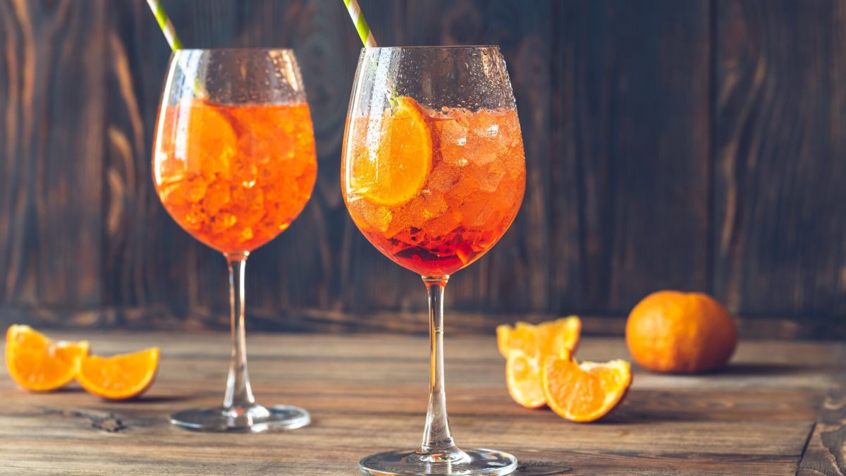 Zwei Gläser mit Aperol Spritz auf hellem Holzuntegrund. Dabeben liegen angeschnittene Orangen.