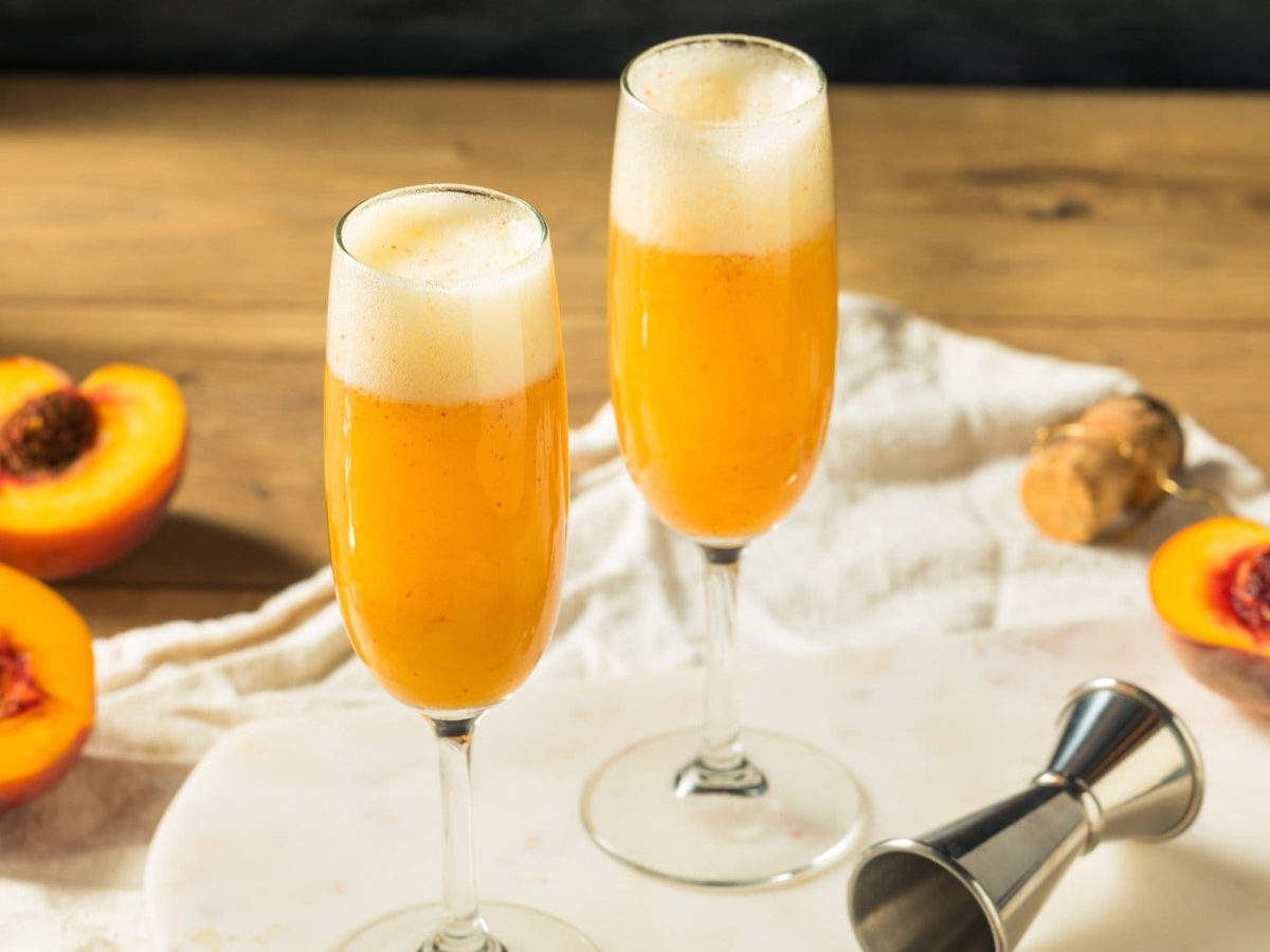 Zwei Gläser gefüllt mit Bellini auf hölzernem Untergrund. Daneben liegen aufgeschnittene Pfirsiche.