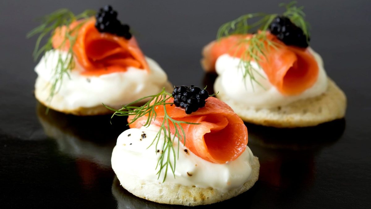 Blini mit Lachs, Sauerrahm und Kaviar dekoriert mit frischem Dill auf glänzendem, dunklen Untergrund.
