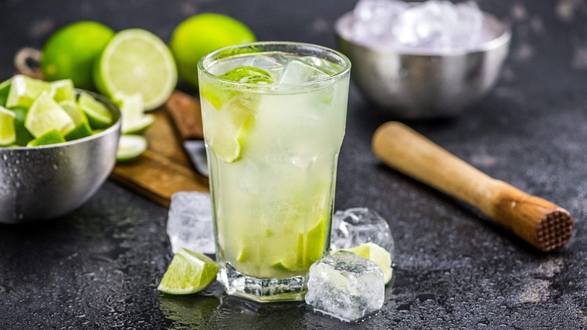 Ein Glas Caipirinha auf dunklem Untergrund. Daneben steht eine Schale mit Limettenwürfeln und eine mit Eiswürfeln. Außerdem ein Brett mit Limetten sowie ein Stößel