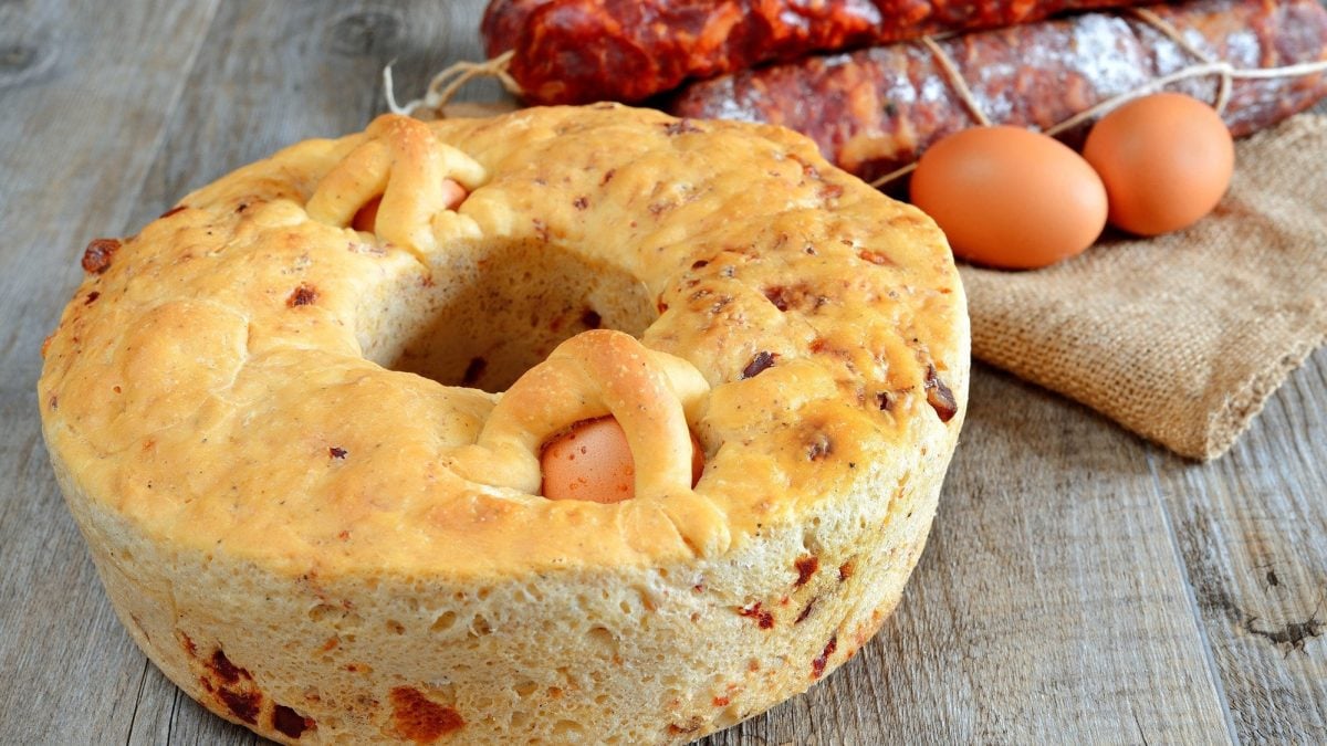 Ein mit hart gekochten Eiern gefüllter, herzhafter Osterkranz aus Italien auf hölzernem Untergrund. Daneben liegen Zutaten wie Eier und Salami.