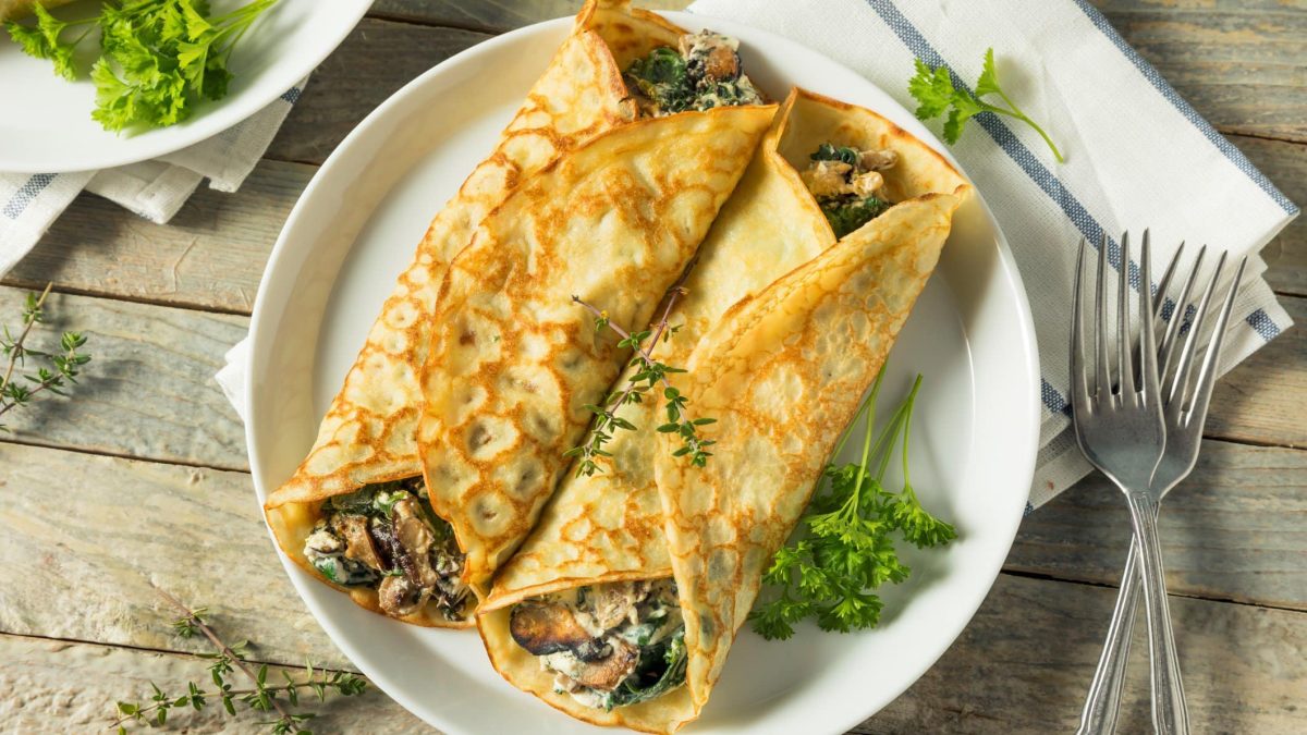 Eingeklappte Crêpes mit Spinat-Pilz-Füllung auf weißem Teller. Dieser steht auf hölzernem Untergrund, daneben liegen zwei Gabeln.