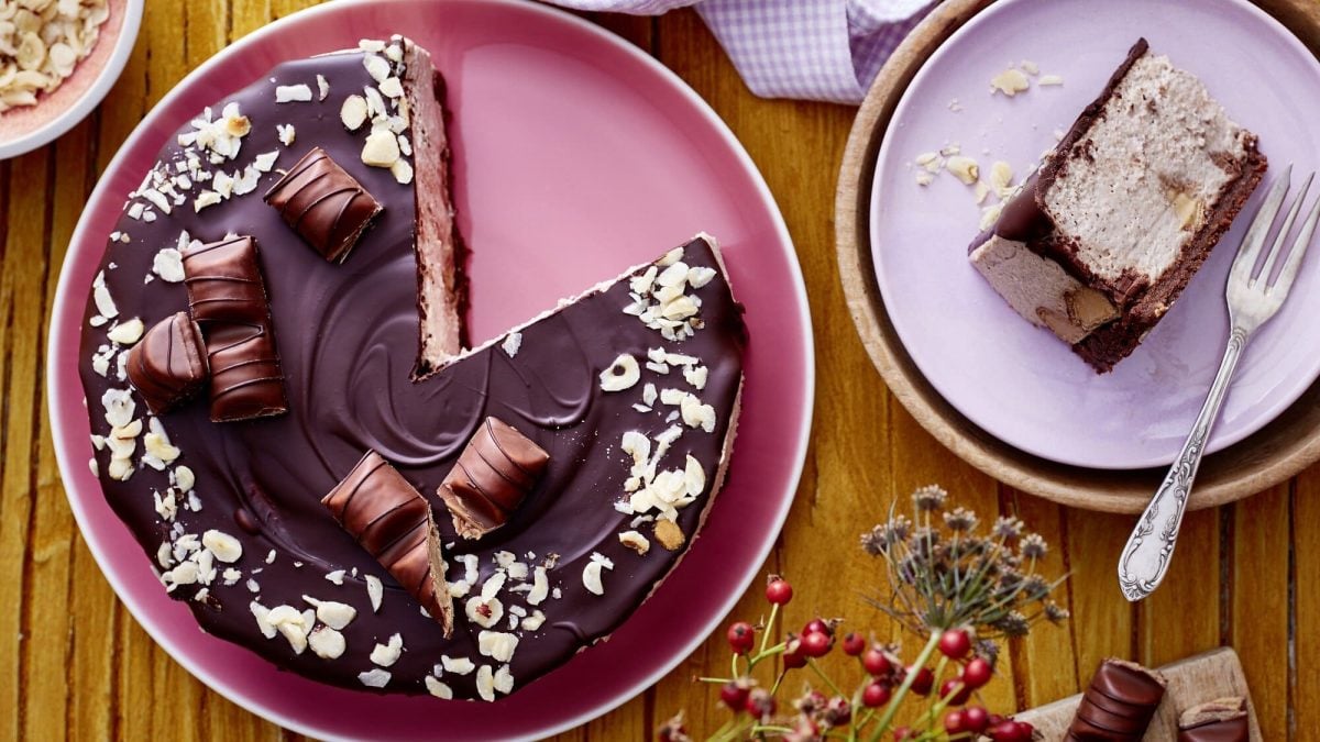 Auf einem rosa Teller liegt ein angeschnittener Haselnuss-Cheesecake. Man erkennt die Creme Schicht in der Mitte. Er ist mit einer Schokoladenglasur überzogen und mit Haselnüssen bestreut.