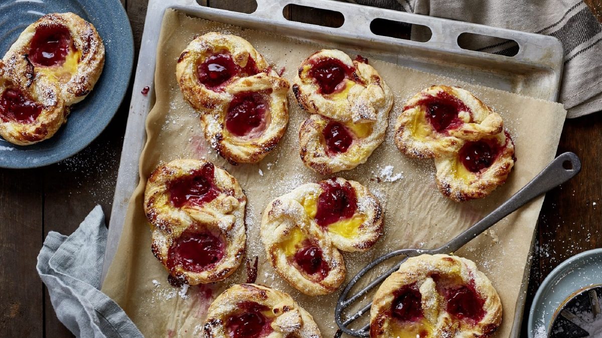 Auf einem Backblech liegen mehrere Pudding-Himbeer-Brezeln. Sie sind aus Blätterteig gemacht und mit Vanillepudding und Himbeerkonfitüre gefüllt.