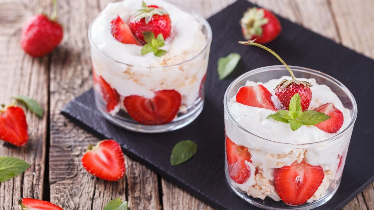 Zwei Gläser mit Eton Mess auf Schieferplatte auf Holzuntergrund. Darum herum liegen Erdbeeren.
