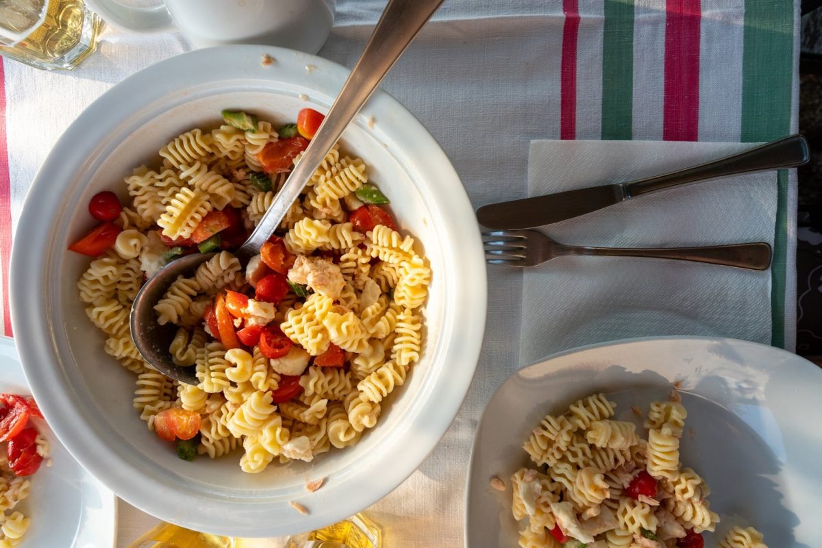 Schüssel mit Nudelsalat auf gemustertem Tischtuch. Daneben steht ein Teller mit Nudelsalat und auch Besteck liegt daneben.
