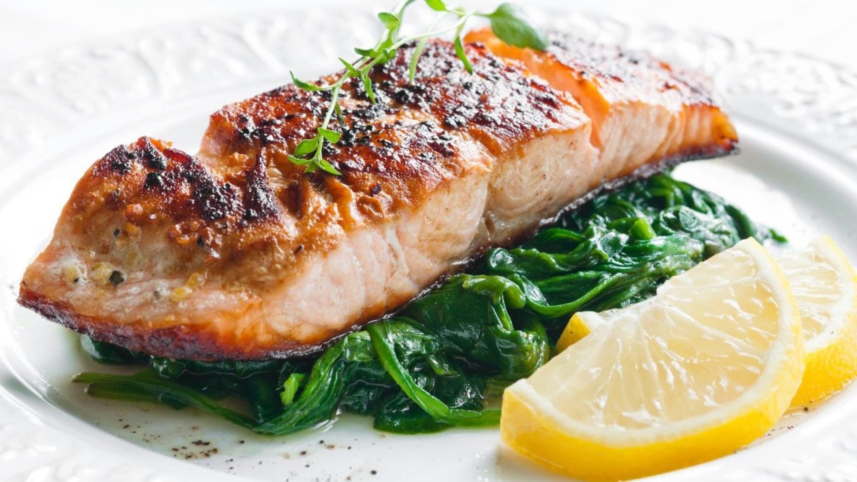 Ein Stück gegrillter Lachs auf Blattspinat auf weißem Teller. Neben liegen zwei Zitronenscheiben.