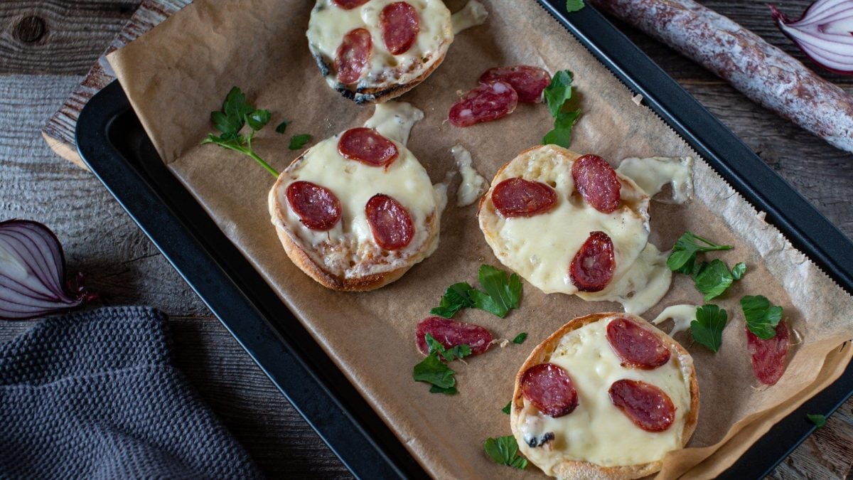 Pizza-Brötchen belegt mit Käse und Salami auf einem mit Backpapier ausgelegtem Blech.