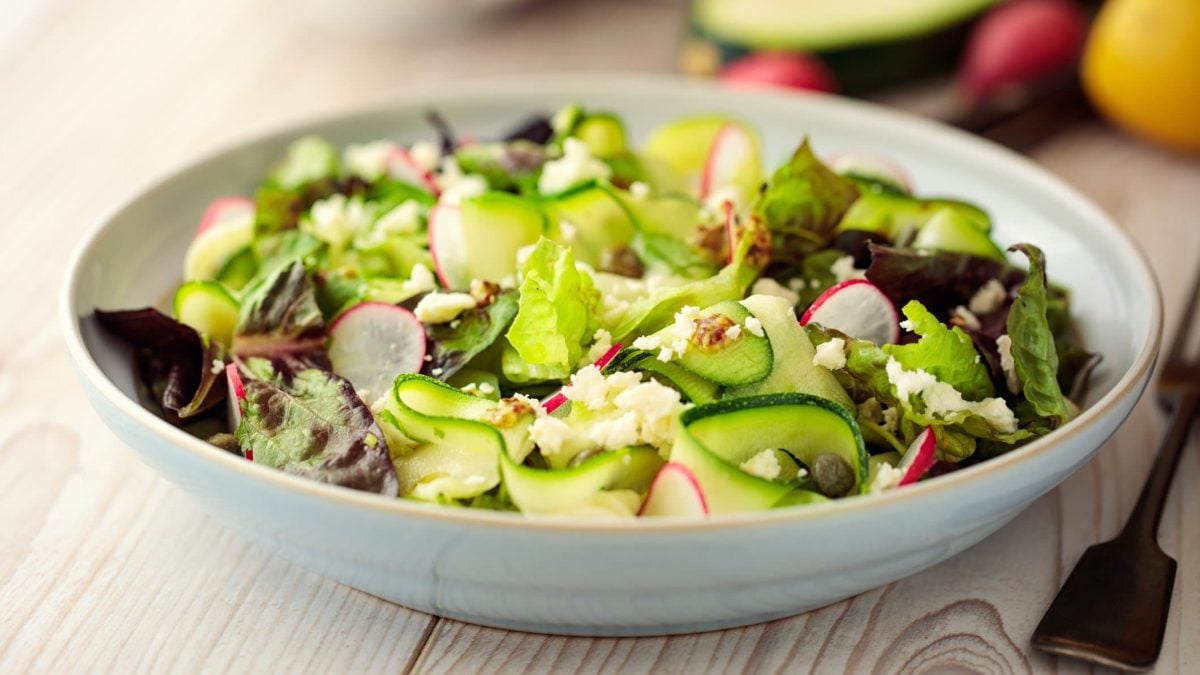 Weißer Teller mit rohem Zucchinisalat, Blattsalat und Radieschen.