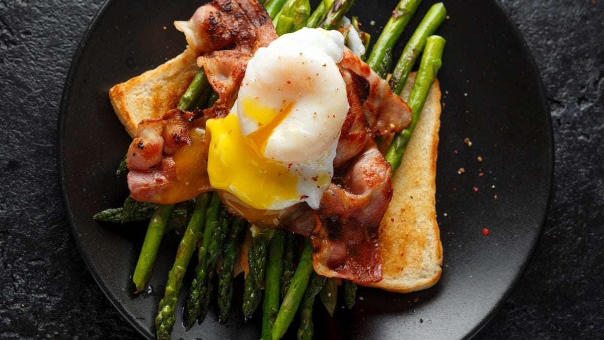 Eggs Benedict mit Spargel und Speck auf einem schwarzen Teller. Dieser steht auf dunklem untergrund.