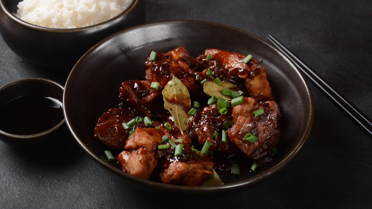 Eine schwarze Schale mit Adobo und Reis im Hintergrund auf schwarzem Tisch.