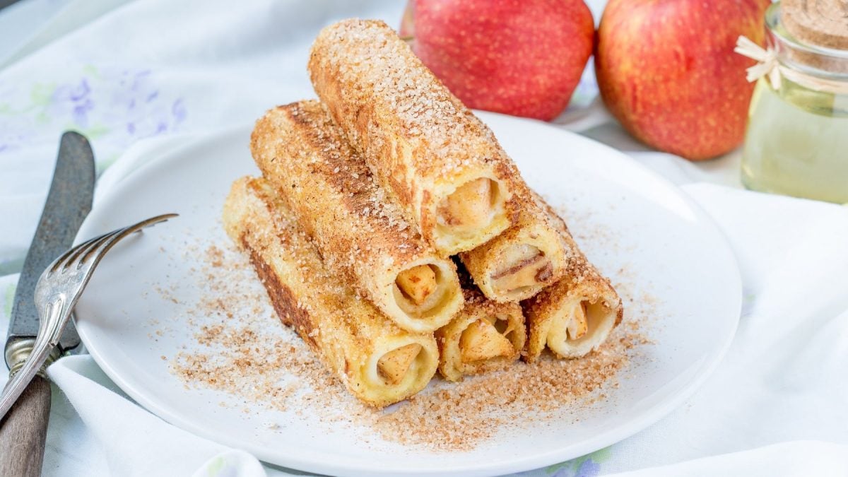 Apfel-Toast-Rollen auf einem weißen Teller. Daneben Besteck und verschiedene Zutaten.