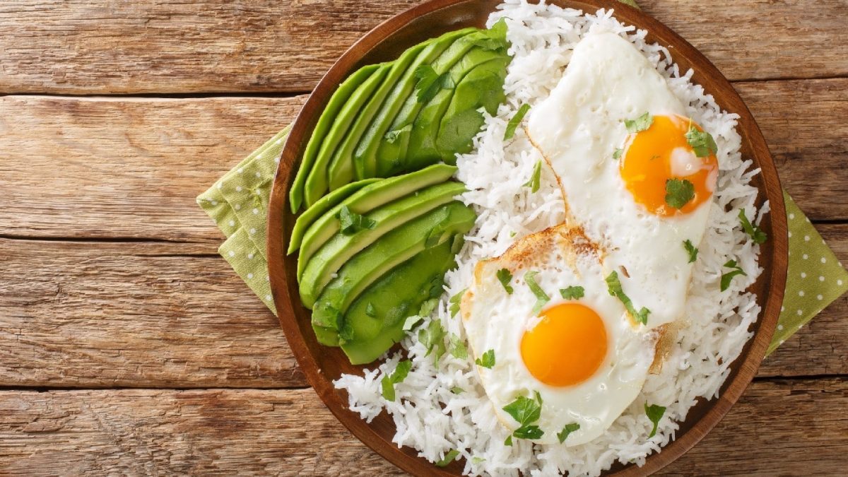 Arroz con huevo frito: Weißer Reis mit zwei Spiegeleiern und Avocado auf einem hölzernen Hintergrund