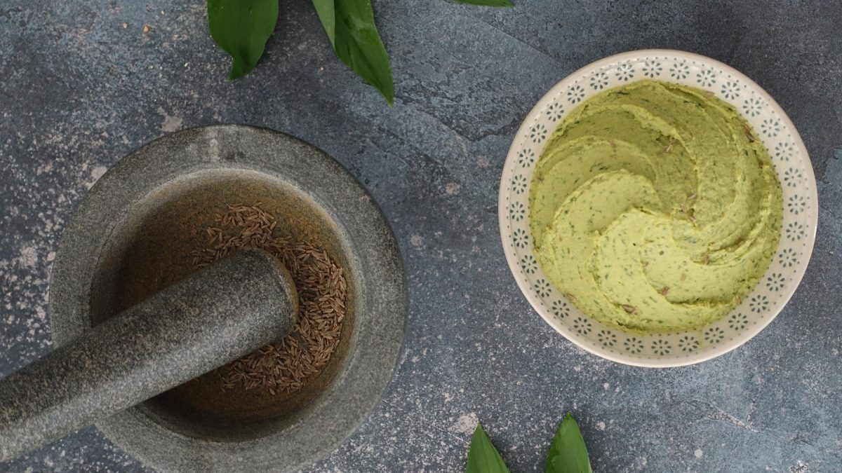 Eine Schale mit Bärlauch-Hummus in der Draufsicht, daneben ein Mörser mit Kümmel und einige Bärlauchblätter.