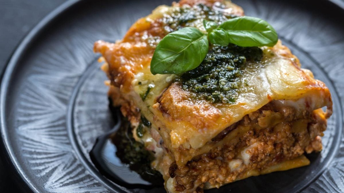 Bärlauch-Lasagne mit selbst gemachtem Bärlauchpesto auf grauem Teller mit Ornamenten auf grauem Untergrund. Basilikumblatt als Dekoration. Draufsicht.