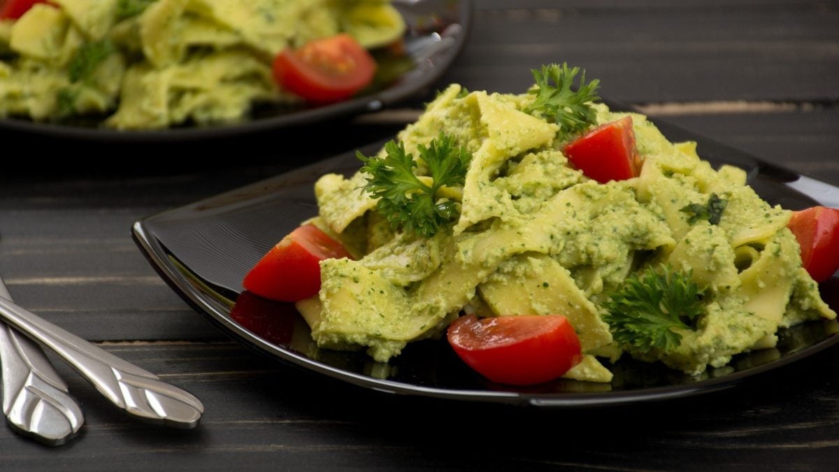 Zwei Teller Bandnudeln mit Cashew-Spinat-Sauce, Tomaten und Petersilie auf dunklem Holz mit Besteck.