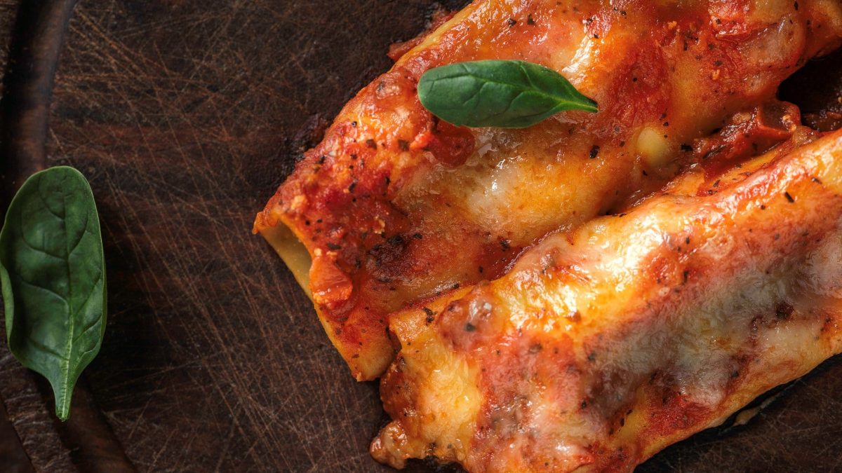 Cannelloni mit cremiger Kalb-Ricotta-Füllung und Bärlauch in fruchtiger Tomatensauce auf dunklem Untergrund mit Bärlauchgarnitur. Aufnahme aus der Vogelperspektive.