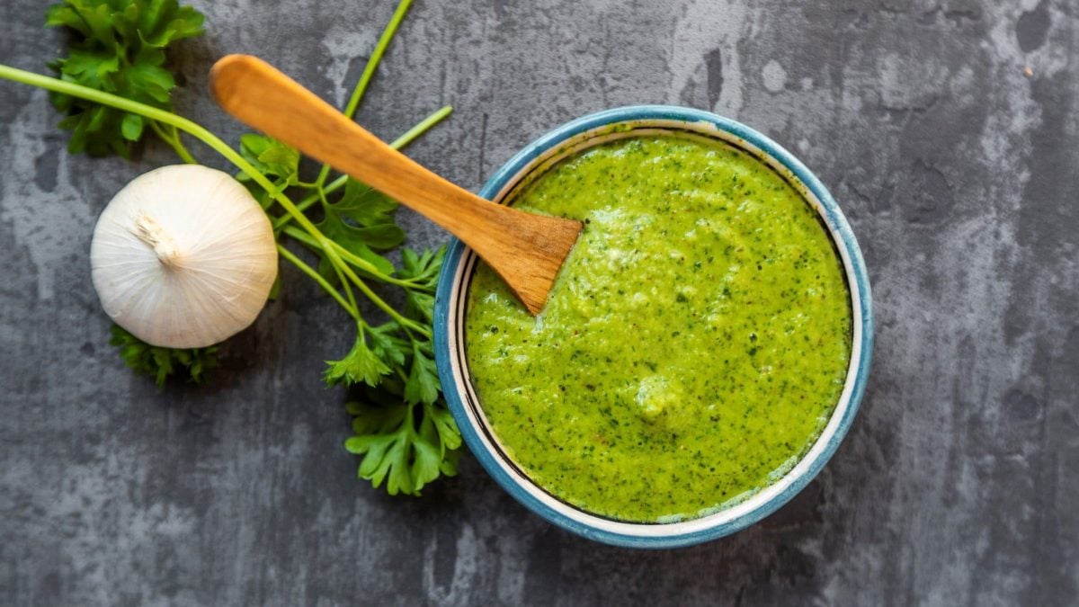 Eine hellblaue Schale mit Chimichurri auf grauem Untergrund. Links davon Petersilie und eine Knoblauchknolle.
