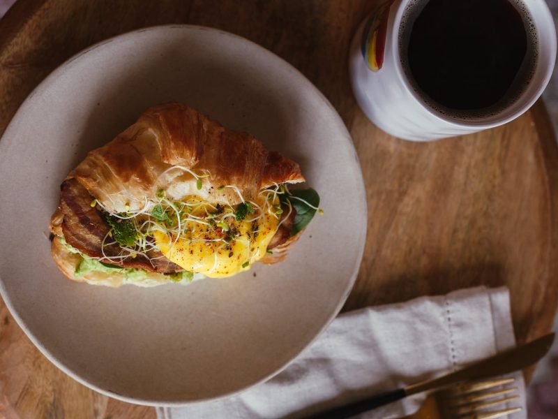 Ein belegtes Croissant mit Ei und Bacon auf einem beigen Teller. Daneben eine Tasse Kaffee und Besteck. Das Ganze auf einem kleinen Holztisch.