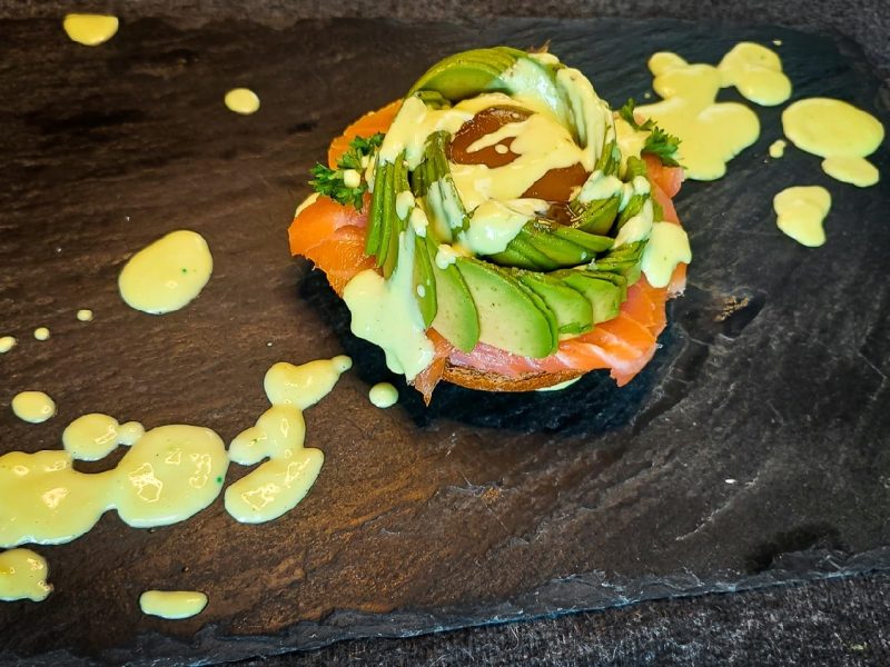 Schwarzer Teller mit dem Egg Benedict in einer Avocasorose auf Lachs und Brötchen. darüber Wasabihollandaise gezogen. Alles auf einer schwarzen Schieferplatte.