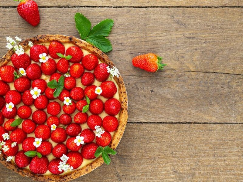 Auf einem Untergrund aus Holz steht ein runder Erdbeer-Vanille-Kuchen mit kandierten Blüten. Um ihn herum liegen Erdbeeren und Erdbeerblätter.