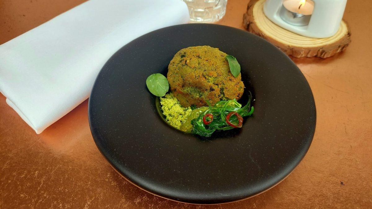 Grün paniertes Falafel-Ei mit Algensalat in einem schwarzen Teller auf einem braunen Tisch.