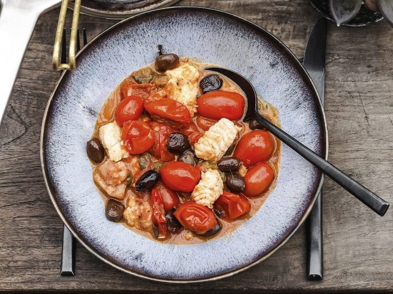 Auf einem Holztisch stehten eine Pfanne und ein Kermaikteller. Auf dem Keramikteller liegt ein Essen: Es ist Fisch mit Gemüse alla Puttanesca