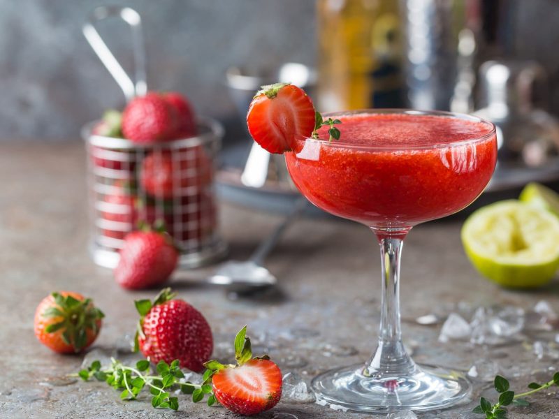 Frozen Strawberry Margarita in Martini-Glas auf braunem Untergrund vor grauem Hintergrund. Dahinter unscharf Erdbeeren im Metallkorb und Cocktail-Zubehör. Auf dem Untergrund Limetten, Erdbeeren und Thymian verteilt. Frontalansicht.
