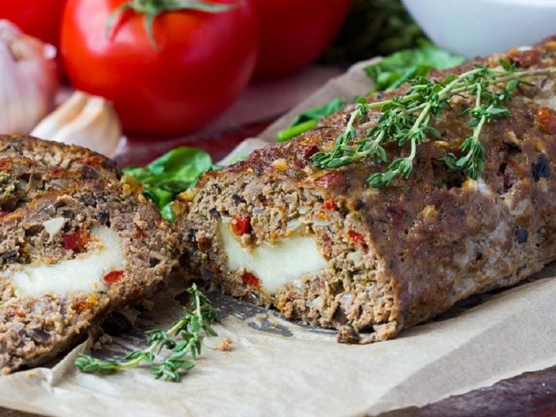 Gefüllter Hackbraten mit Käse und Schmorgemüse zum Muttertag auf weißem Teller mit frischem Thymian. Im Hintergrund Tomaten und Knoblauch. Frontalansicht.
