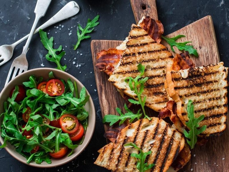 auf einem Holzbrettchen liegt ein gegrilltes Weißbrot mit Speck und Mozzarella. Daneben liegen noch zwei weitere Sandwiches. Und auch noch eine Schüssel mit einem Rucola-TOmaten-Salat.