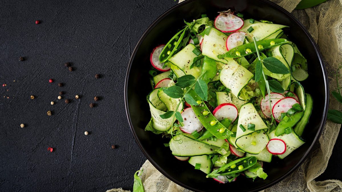 Ein dunkler Teller Gurkensalat mit Zuckerschoten, Radieschen und frischen Kräutern auf dunklem Untergrund in der Draufsicht.