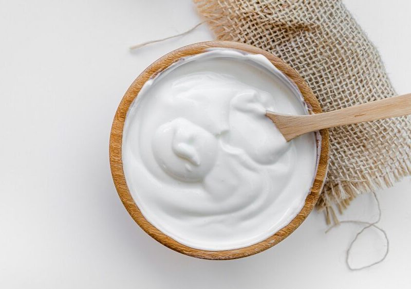 Aus der Vogelperspektive. In einer Schüssel aus Holz ist einen weiße Creme. Es handelt sich um selbstgemachten Joghurt. In dem Joghurt steckt ein Löffel aus Holz.