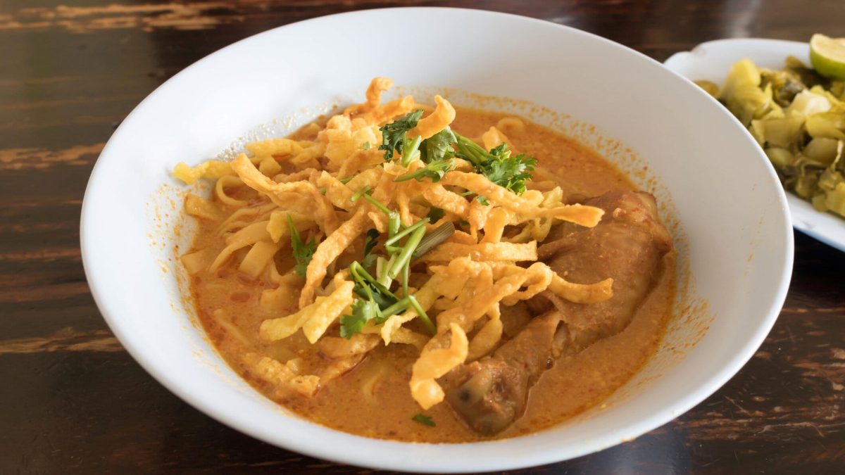 Eine weiße Schale Khao Soi in der Draufsicht auf einem Holztisch.