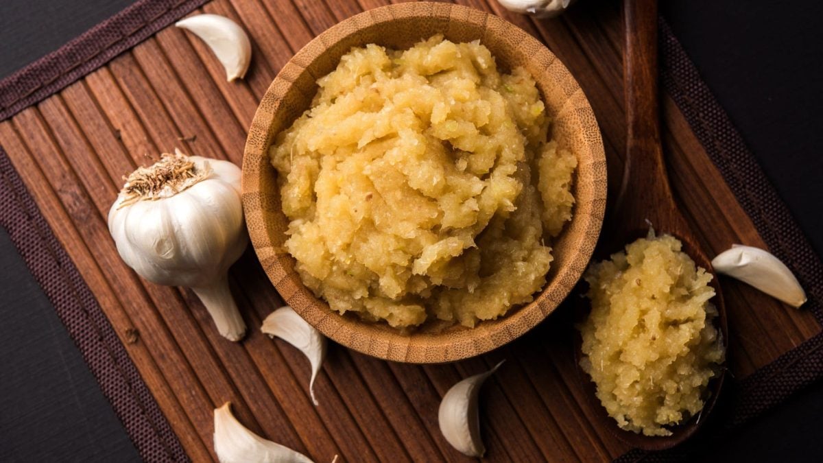 Geröstete Knoblauchpaste auf Holzbrett auf dunklem Untergrund. Daneben Holzlöffel mit der Paste und Knoblauchzehen verteilt. Aufnahme aus der Vogelperspektive.