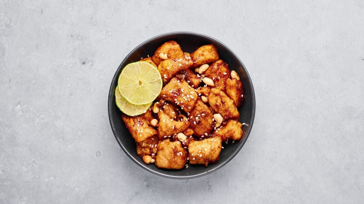 Koranisches Dakgangjeong Korean Fried Chicken in einer schwarzen Schale mit Sesam, Erdnüssen und Zitronenscheiben garniert auf grauem Untergrund. Aufnahme aus der Vogelperspektive.