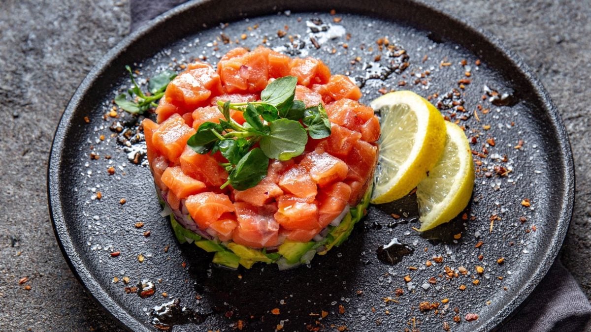 Lachstatar auf Avocado angerichtet in einem Ring auf einem schwarzen Teller. Mit vielen Gewürzen drum herum.