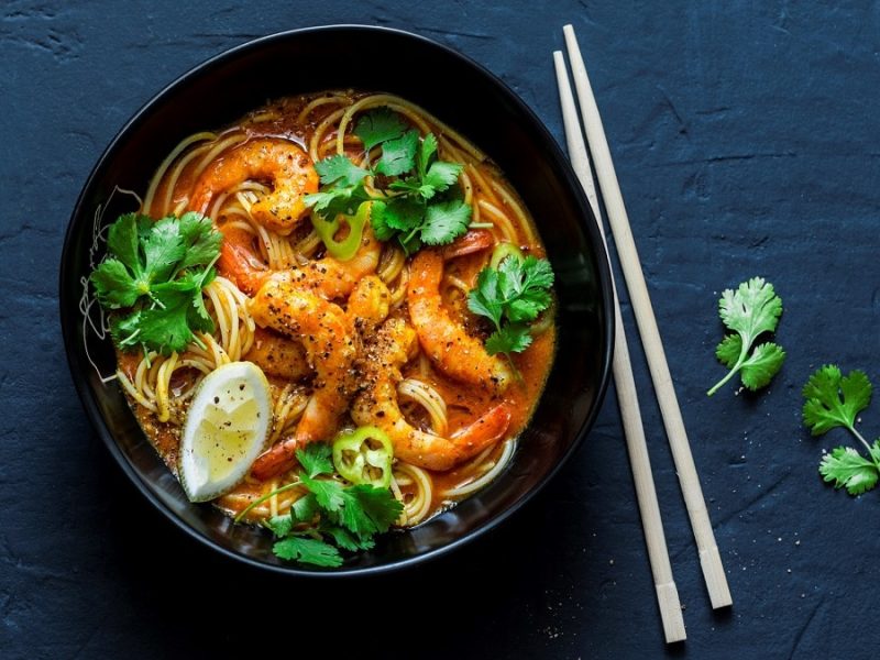 Laksa-Suppe mit Garnelen mit Koriander und einer Scheibe Zitronen in einer schwarzen Schüssel mit einem Paar Stäbchen auf einem schwarzen Hintergrund