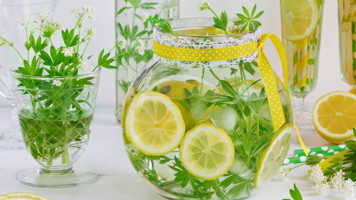 Eine Karaffe Maibowle mit frischem Waldmeister und Zitronenscheiben, daneben Gläser mit Bowle und Waldmeister.